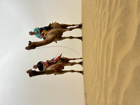       Two travellers riding camels across rippled golden sand dunes under an overcast desert sky.
  