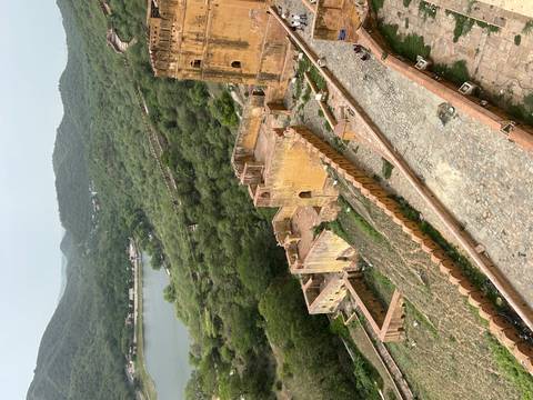       Elevated view of ancient fort walls and yellow sandstone buildings surrounded by green hills and a lake.
  