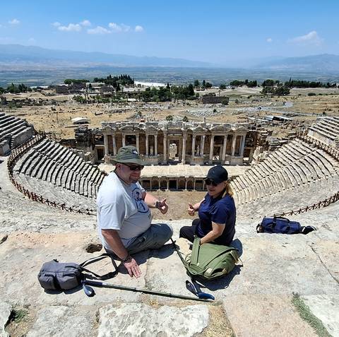       Two tourists sit on the ancient stone tiers of Hierapolis theatre overlooking the Roman stage and surrounding ruins in Pamukkale.
  