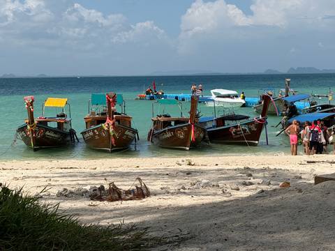       Four colorful long-tail boats tied up along a white-sand beach with turquoise water and distant limestone islands.
  