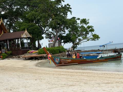       A decorated long-tail boat with tourists approaches a small beachside restaurant among lush trees.
  