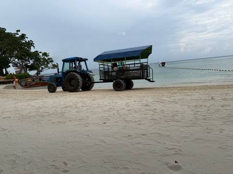       A blue tractor pulling an open passenger cart sits on a wide sandy beach near calm water.
  