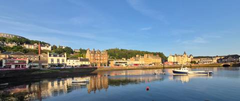       Colorful waterfront buildings of Oban reflected crisply in calm harbor waters at golden hour.
  