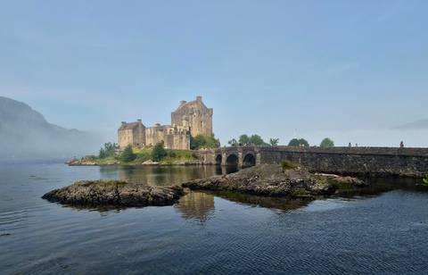       Eilean Donan Castle rises from mist above a stone bridge and reflective tidal waters.
  