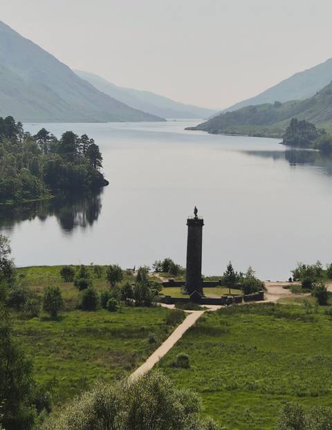       Tall stone Glenfinnan Monument stands beside a tranquil loch surrounded by forested hills.
  