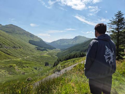       Hiker in hoodie surveys a long valley of dense forest and rolling hills beneath blue skies.
  