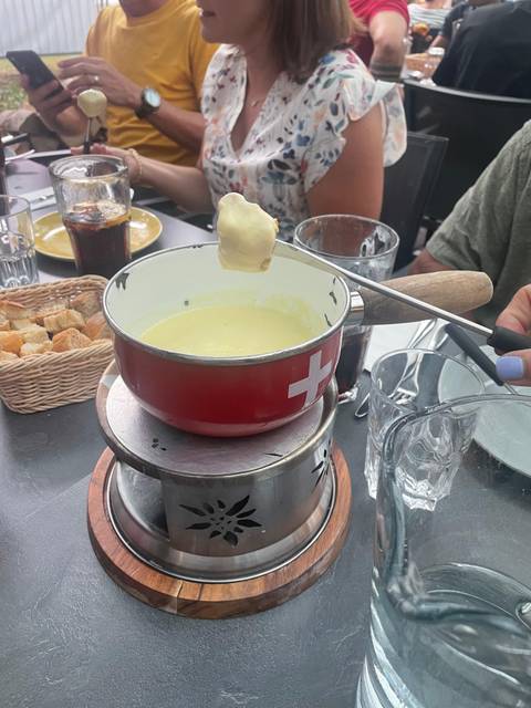       A red fondue pot filled with melted cheese sits on a burner surrounded by cubes of bread and drinks on an outdoor restaurant table.
  