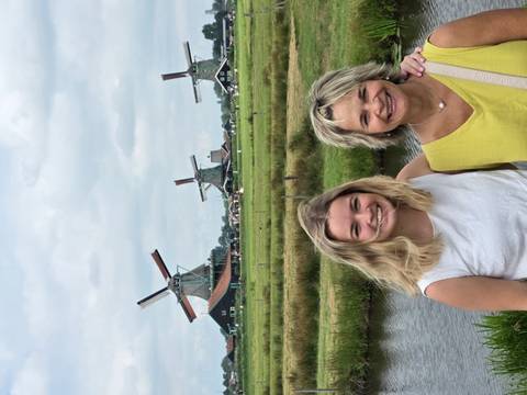       Two smiling women pose in front of classic Dutch windmills amid green fields and a small canal under a cloudy sky.
  