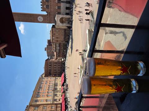       Two tall glasses of beer rest on a balcony railing overlooking a sunny Italian piazza filled with historic brick buildings.
  