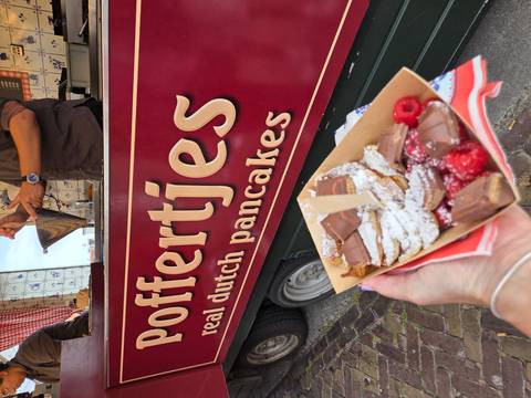       A tray of freshly cooked poffertjes dusted with powdered sugar is held up in front of a maroon Dutch pancake food truck.
  