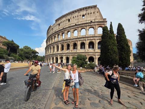       Crowds gather outside the Colosseum while two cheerful women pose in front pointing toward the ancient arena.
  