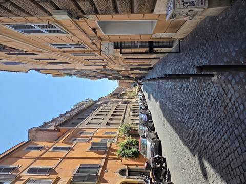       A long, narrow cobblestone street lined with colorful Roman apartment buildings stretches into the distance under a blue sky.
  