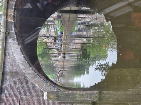       A view through a brick arch bridge reflects along an Amsterdam canal with trees and houses lining the water.
  