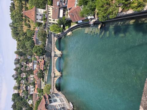       A clear turquoise river flows beneath a stone bridge toward a hillside town of chalet-style homes and trees.
  