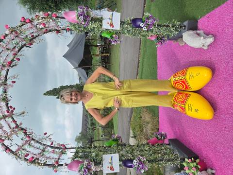      A woman stands on bright pink carpet wearing enormous decorative yellow wooden clogs under a floral arch.
  