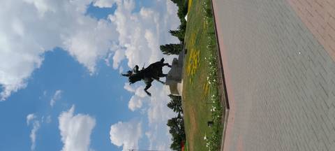       A bronze equestrian statue rises on a stone pedestal in a landscaped park under a partly cloudy sky.
  