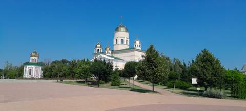       A pale cream Orthodox cathedral with four golden domes stands surrounded by green trees beneath a clear blue sky.
  