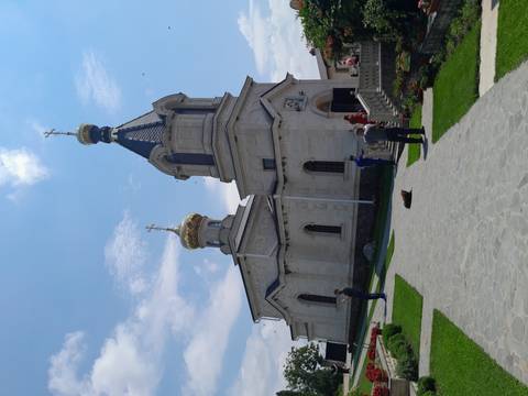       People walking around a white stone Orthodox church with golden onion domes under a bright blue sky, a dog lying on the pathway in front.
  