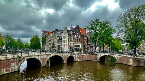       Historic brick bridge spanning an Amsterdam canal lined with narrow gabled houses beneath dramatic storm clouds.
  