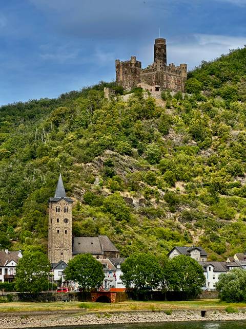       Pointed church steeple peeks above a densely forested rocky hillside under bright daylight.
  