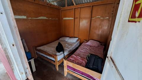       Simple wooden cabin room with two single beds, backpacks and plain brown walls.
  