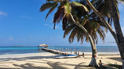       Sandy beach with a long wooden jetty, anchored boat and leaning palm trees in bright sun.
  