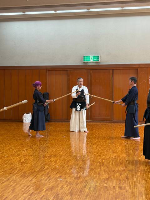       Martial arts instructor demonstrates wooden sword techniques with two students in a polished dojo.
  