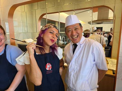       Smiling participant and chef enjoy a sushi-making class, holding chopsticks and dressed in aprons and chef hat.
  