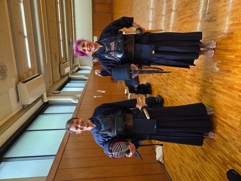       Two travelers wear full kendo armor and hold helmets proudly in an indoor hall.
  