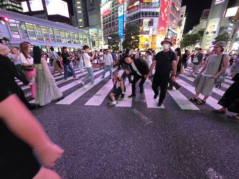       Crowded night scene at Shibuya Crossing with a traveler kneeling on the zebra stripes amid bustling pedestrians.
  