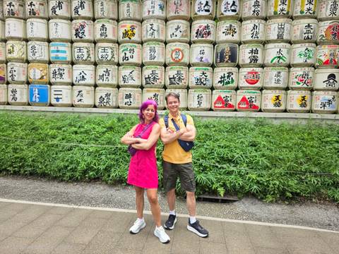       Smiling couple stand before a wall of decorative sake barrels in a leafy shrine setting.
  