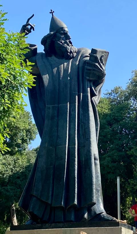       Close-up of a large dark bronze statue against greenery and blue sky, details largely in shadow.
  