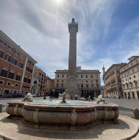       Fountain-centred piazza with a tall ancient column surrounded by classic Roman buildings under a partly cloudy sky.
  