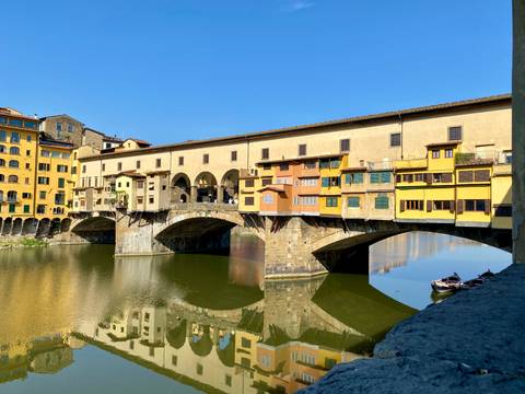       Historic Ponte Vecchio bridge lined with pastel shops spans the calm Arno River in Florence under a clear sky.
  