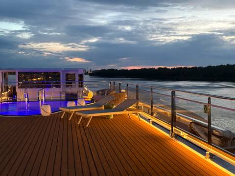       Sunset view from a cruise-ship sundeck with loungers, pool lights and a calm river.
  