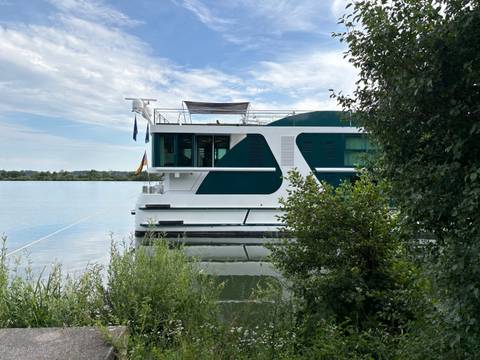       Modern river ship docked beside calm water and green shoreline under a blue sky.
  