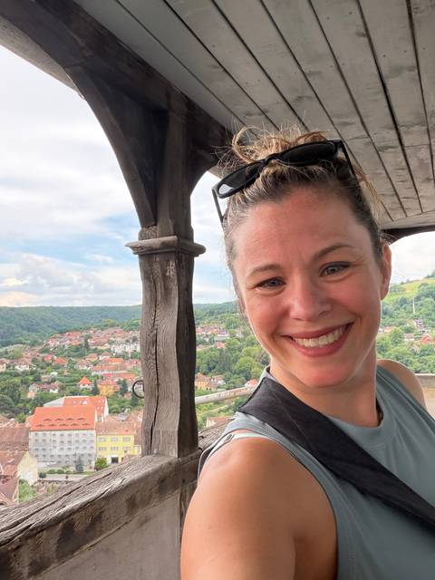       Woman smiling from a wooden lookout with rolling green hills and tiled rooftops in the valley below.
  