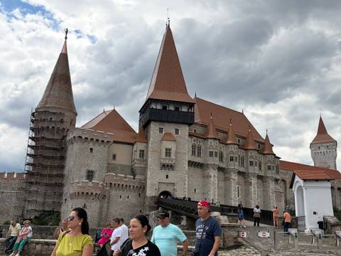       Visitors entering Corvin Castle with its red-tiled roofs and towers under brooding clouds.
  