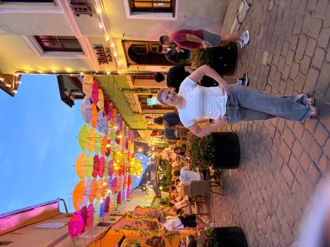       Colorful umbrella-covered alley lit by fairy lights with diners and a posing visitor at dusk.
  