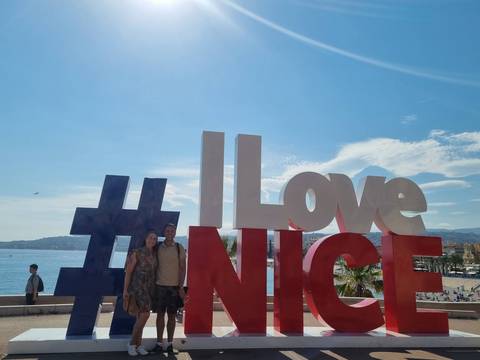       Couple standing beside the huge #ILoveNICE sign on the Nice seafront with blue sky and Mediterranean backdrop
  