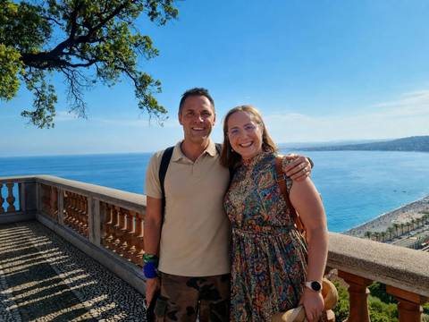       Smiling couple on a balcony overlooking the turquoise bay and shoreline of Nice
  