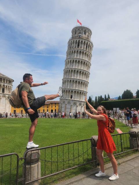       Two travellers doing a playful forced-perspective pose with the Leaning Tower of Pisa on a bright day
  