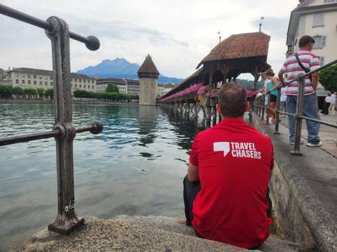       Man in red Travel Chasers shirt sitting by Lake Lucerne looking toward the historic Chapel Bridge and water tower
  