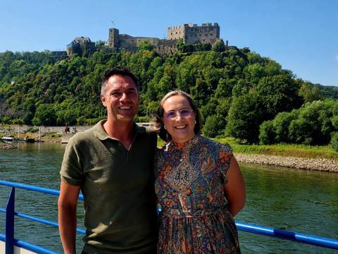       Couple on a river boat with a hill-top Rhine castle and green forested slopes behind
  