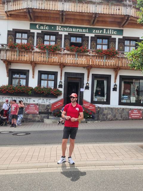       Tour guide in red shirt standing in front of a traditional Black Forest style restaurant and café
  