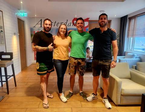       Four travellers posing happily inside a hostel lounge with an Amsterdam poster behind
  