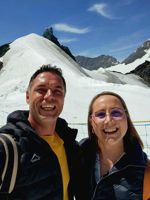       Selfie of two travellers with snowy alpine slopes and glacier behind them
  