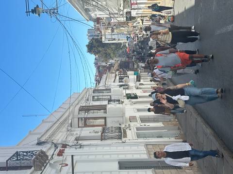       Busy colonial street lined with white-washed buildings as tourists walk uphill beneath a bright blue sky.
  