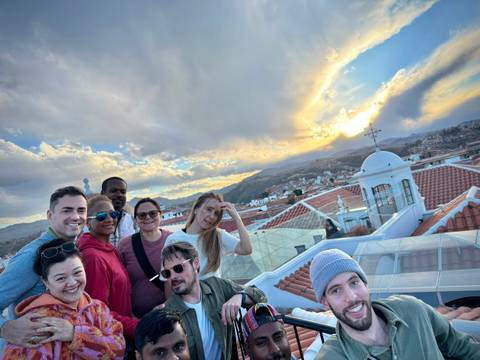       Smiling friends crowd together for a selfie on a rooftop terrace overlooking red-tiled roofs and a dramatic sunset sky.
  