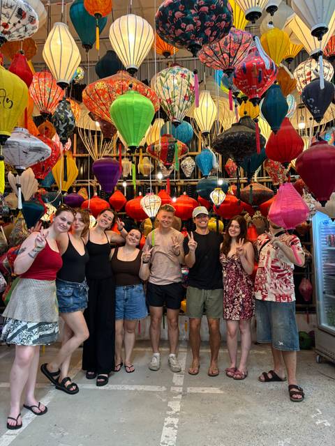       Smiling travellers pose making peace signs amid a vibrant stall filled with colorful silk lanterns.
  